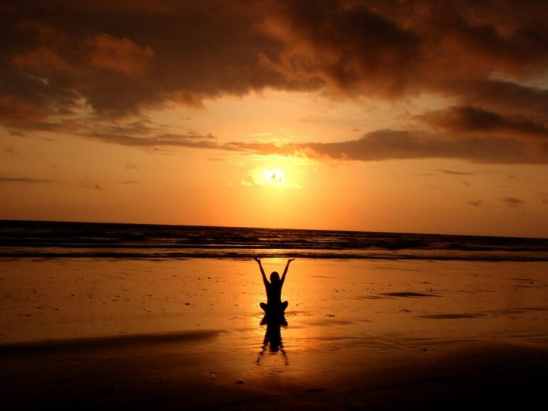 A woman sitting on the beach at sunset, raising her hand, symbolizing deep relaxation, freedom, and connection with nature.