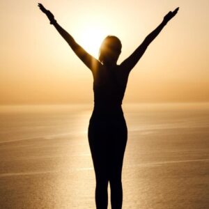 Woman raising her hand on the beach at sunset, symbolizing handling conflict with confidence through hypnosis.