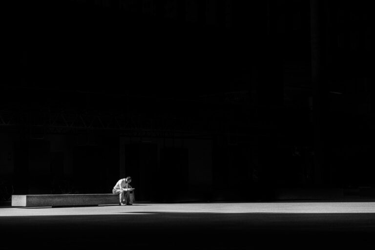 Person sitting alone on a bench with warm light breaking through, symbolizing hope in solitude.