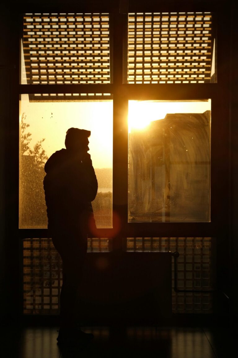A person standing at a window with sunlight streaming in, symbolizing resilience, hope, and inner strength despite challenges.