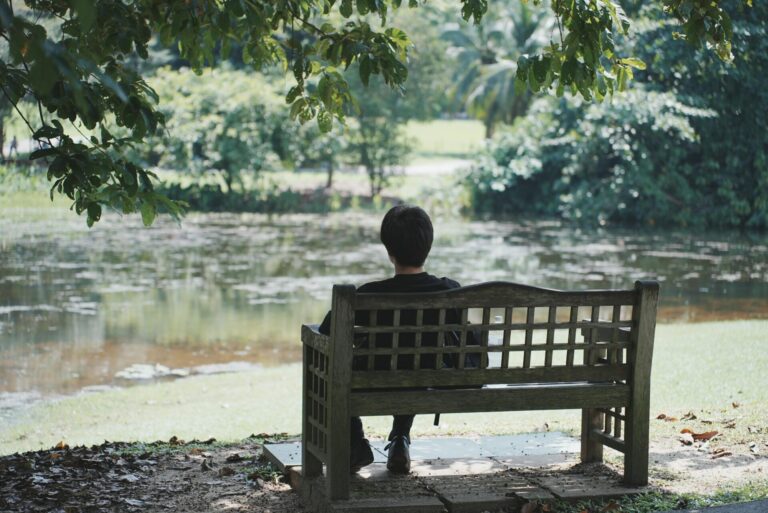 Person sitting cross-legged in front of a calm lake, meditating and enjoying peaceful surroundings.