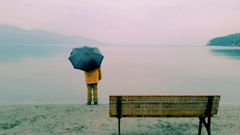 Person standing under an umbrella in gentle rain by the beach, surrounded by a soft glow, symbolizing protection and calm in vulnerable moments.