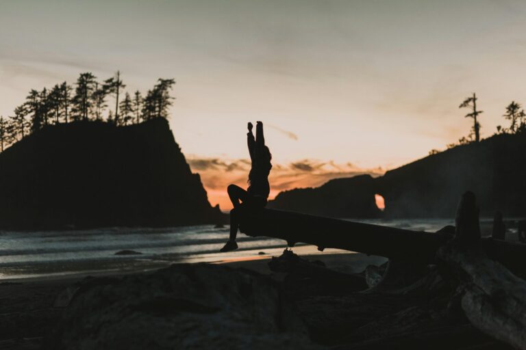 Person sitting cross-legged on a calm beach at sunrise, symbolizing presence and grounding.