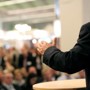 A person in a suit gesturing while speaking at a podium in front of a blurred audience.