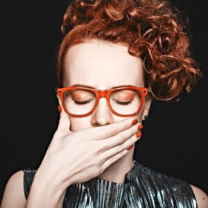 A woman with curly red hair and red glasses covers her mouth with her hand.