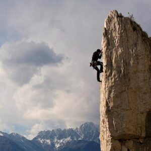 A person climbs a tall, steep rock face with mountains in the background.