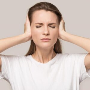 A woman in a white shirt with eyes closed, covering her ears with her hands.