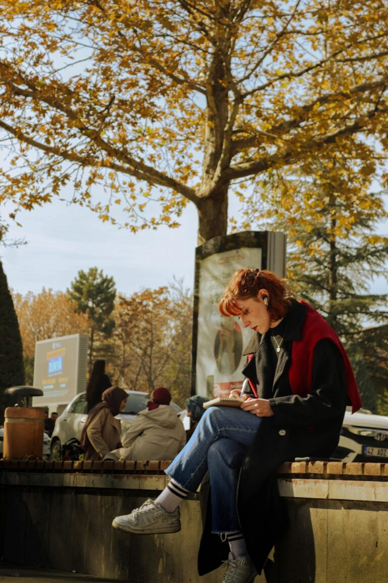 A young woman in a long coat sits on a ledge outdoors, writing in a notebook under an autumn tree.