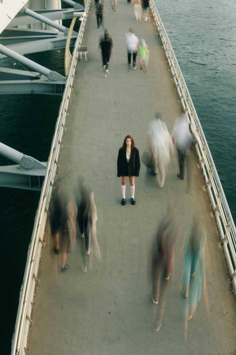 A solitary woman stands still on a busy bridge while blurred figures walk past her, highlighting her isolation in a crowd.