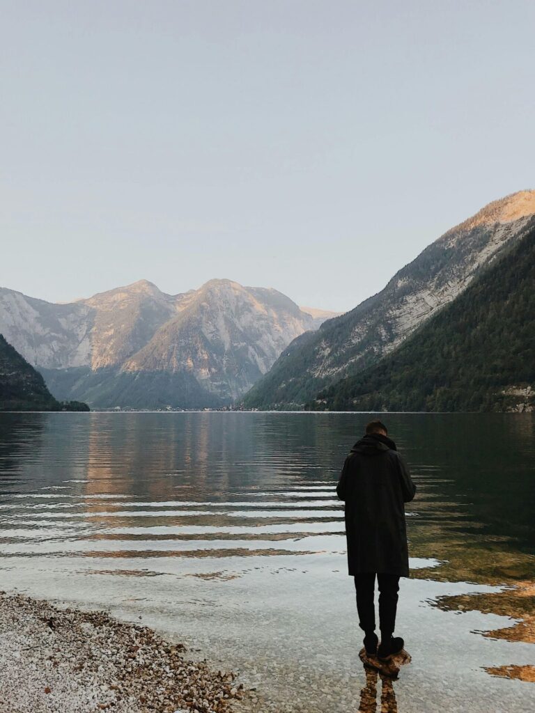 A person in a dark coat stands at the edge of a calm lake, surrounded by mountains and peaceful scenery.