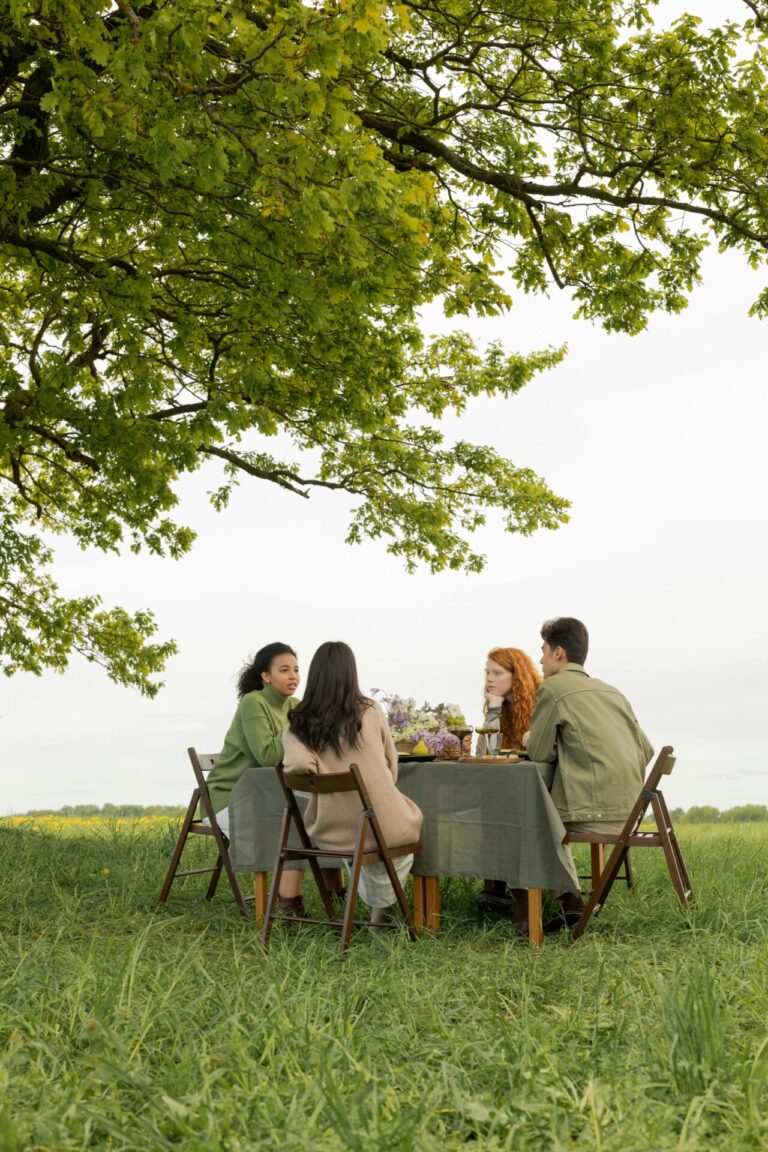 Four friends sit together at a table under a large tree, enjoying a picnic in a green field.