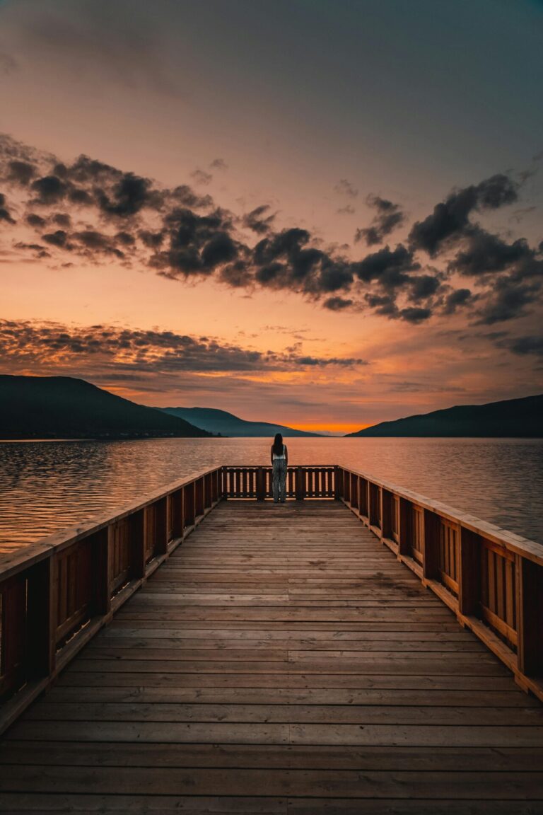 A person stands at the end of a wooden pier, gazing at a colorful sunset over a calm lake surrounded by mountains.