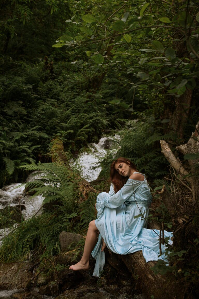 A woman in a flowing light blue dress sits on a log beside a forest stream, surrounded by lush greenery.