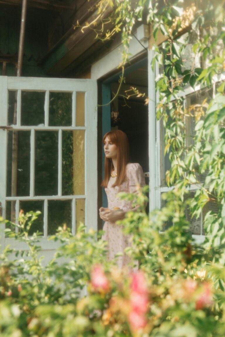 Young woman in a light pink dress stands at an open door, surrounded by lush garden greenery and flowers.