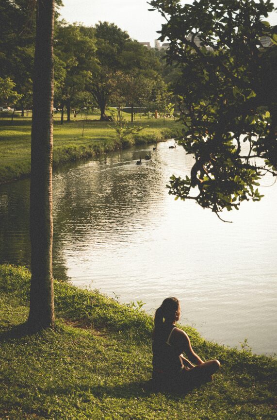 A woman sits cross-legged on the grass by a peaceful lake, enjoying the tranquil natural surroundings.