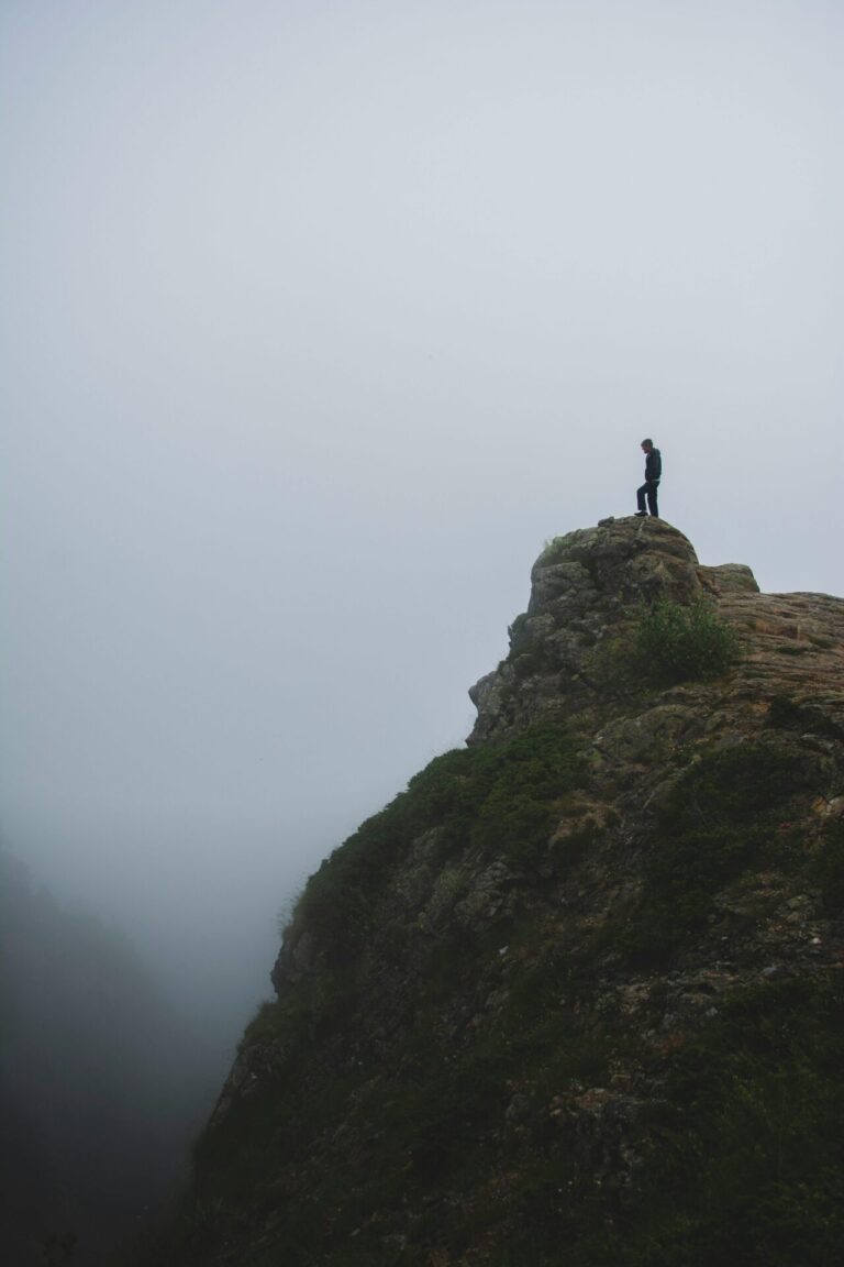 A person sits peacefully on a mountaintop, surrounded by panoramic views of distant peaks under a serene sky.