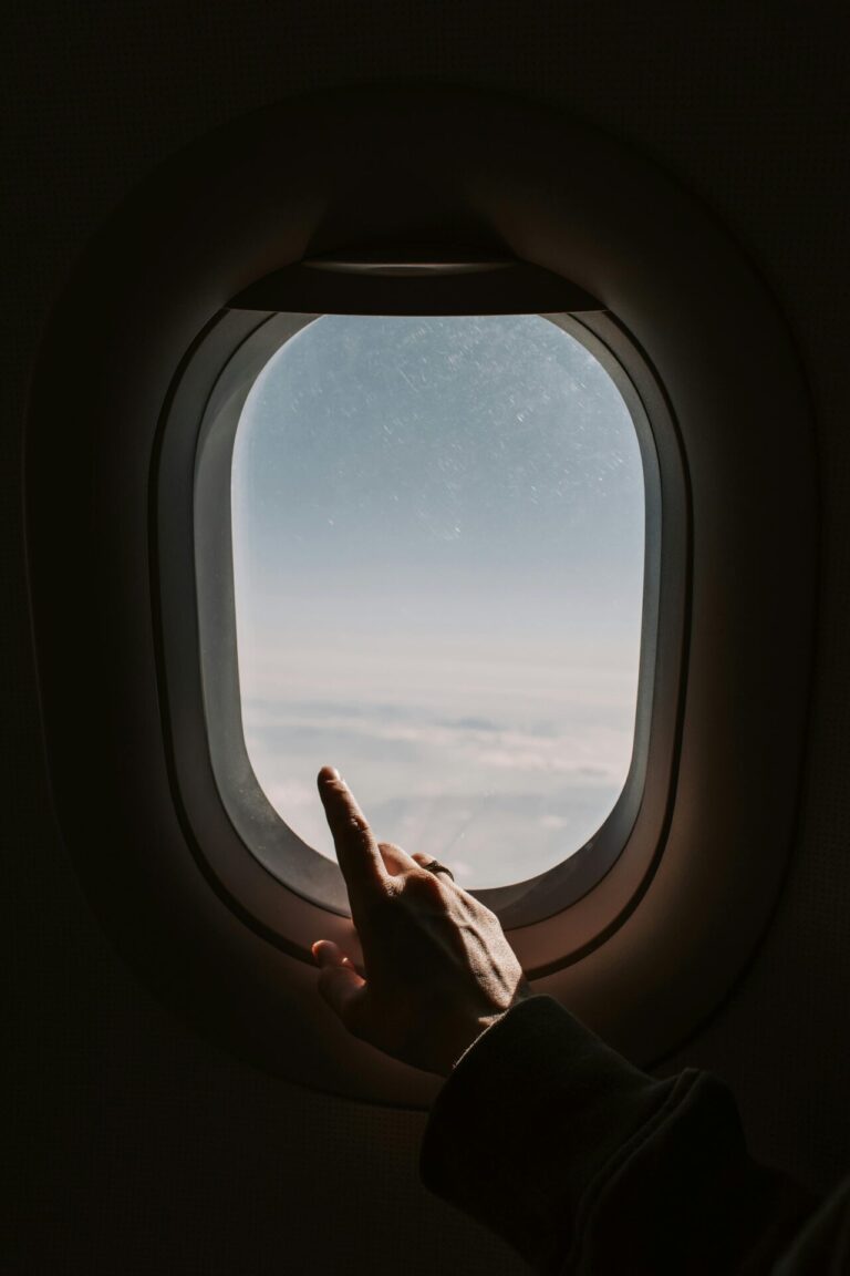 A hand points toward the clouds outside an airplane window during a flight.